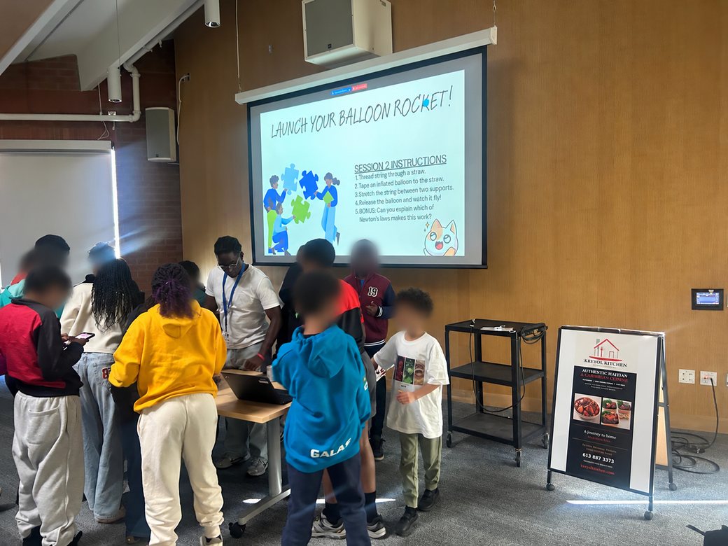 Students gather around a table to build balloon rockets, beside a projected slide titled "Launch your balloon rocket!" with step-by-step instructions.