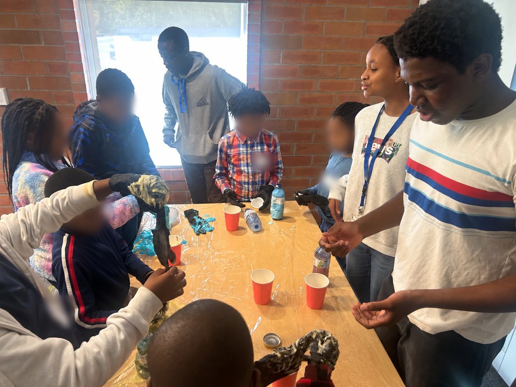 Youth volunteers wearing lanyards guide young learners through a hands-on STEM activity with gloves, cups and water bottles at a table.